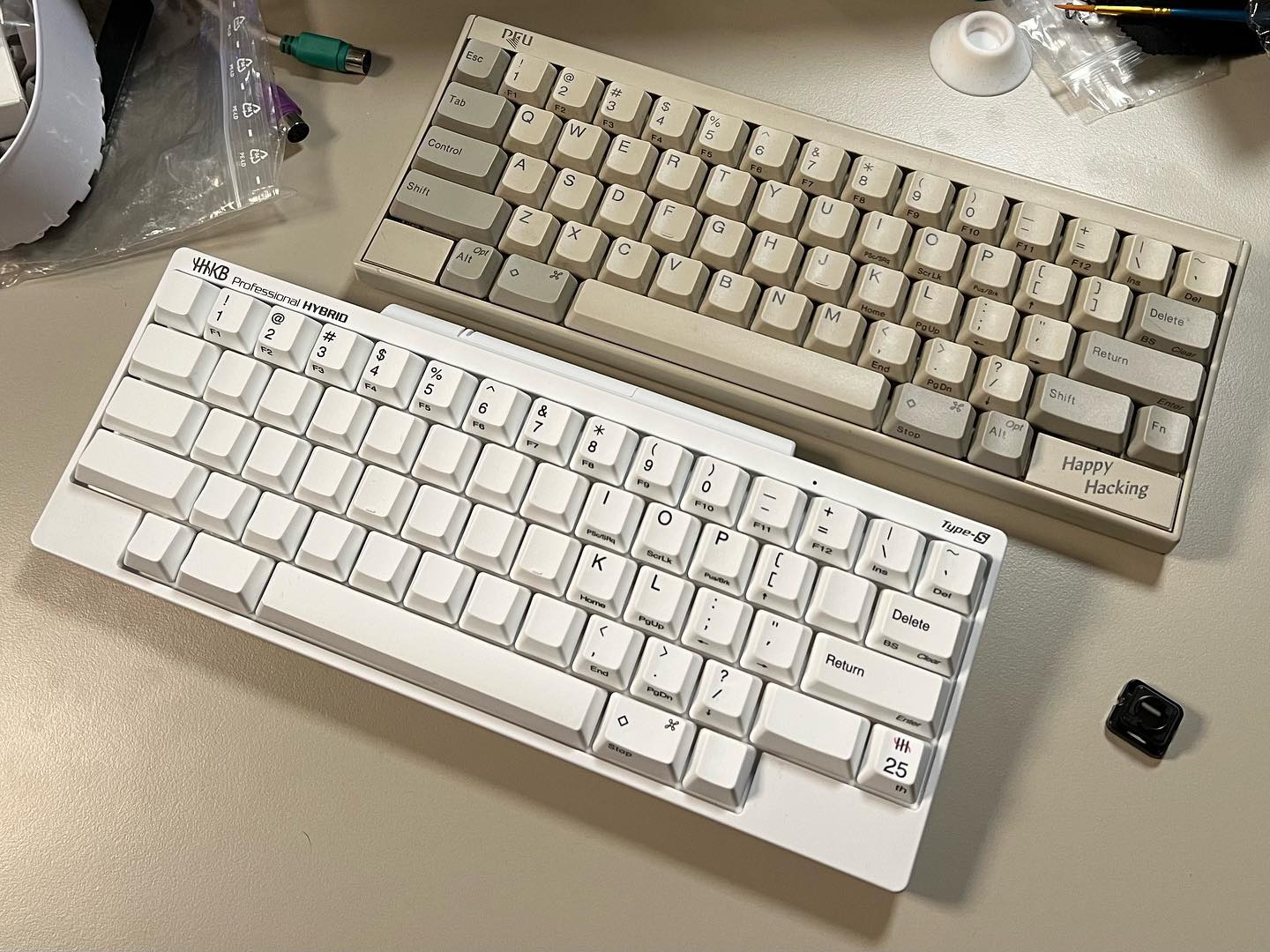 photograph of a Happy Hacking Keyboard 2 shown sitting on a desk
        just above a HHKB Professional HYBRID Type-S Snow model released for the
        original HHKB's 25th anniversary, the latter of which has some of its
        keycaps replaced with blank ones to highlight the special Fn layer
        functions on the remaining keycaps, which are those inherited from the
        original model